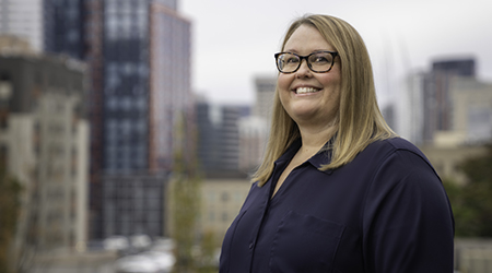 Denise Kirby, a woman in a blue shirt standing in front of a city.